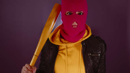 Young woman in pink balaclava with baseball bat. Hooligan in mask scratching head with bat and...