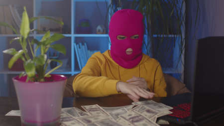Young woman in pink balaclava collects money from table in office. Hacker in mask with dollar bills...