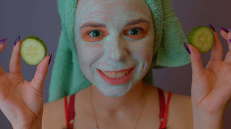 Young woman with towel on head and mask on face hiding her eyes behind cucumbers and showing them....