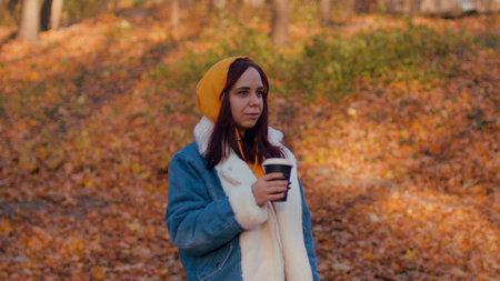 Young woman drinking coffee in autumn forest. Beautiful female in casual clothes enjoying hot drink in paper cup standing in sunny seasonal forest.の写真素材