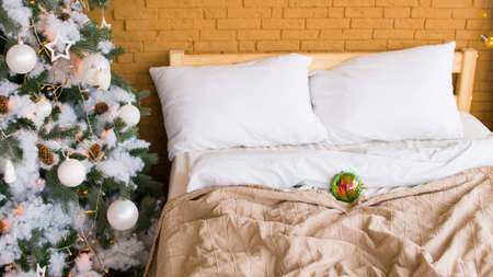 Christmas tree with different christmas toys standing near bed in room. Decorated coniferous tree with white and silver adornments to create festive mood during holidays indoors.の写真素材