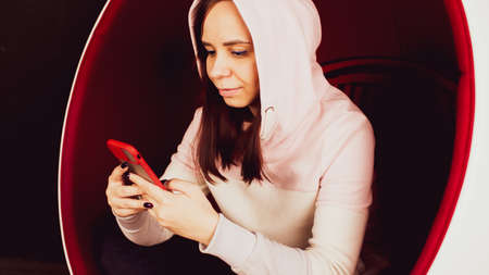 Young woman with mobile phone sitting in ball chair on black background. Female browsing smartphone and resting in modern armchair.の写真素材