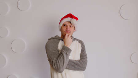 Man in santa hat scratching chin on background of white patterned wall. Thoughtful male looking away and reflecting about upcoming holidays.の写真素材