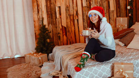 Smiling woman with Christmas gift. Young smiling female in Santa hat warm clothes and eyeglasses sitting on bed with wrapped Christmas gift box in studioの写真素材