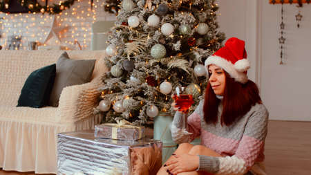 Young woman in santa hat sitting on floor surrounded by gift boxes and Christmas tree. Positive female drinking alcohol and celebrating New Year in festive atmosphere.の写真素材