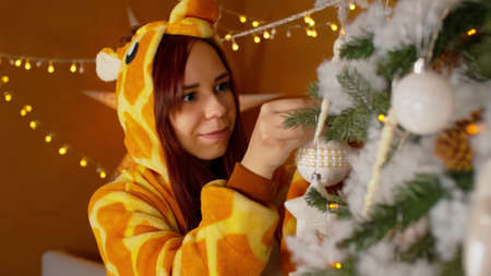 Woman in costume of giraffe decorating and hanging baubles on Christmas tree. Female in funny pyjamas of giraffe decorating a Christmas tree.の写真素材