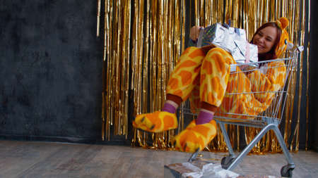 Smiling in kigurumi with Christmas presents in trolley. Side view of excited young female in kigurumi pajama sitting in trolley with Christmas gifts in shiny wrapper against wall with golden tinselの写真素材