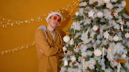 Young man in santa hat standing with arms crossed near Christmas tree and looking at camera. Handsome male in fashionable glasses smiles, posing near decorated tree.の写真素材