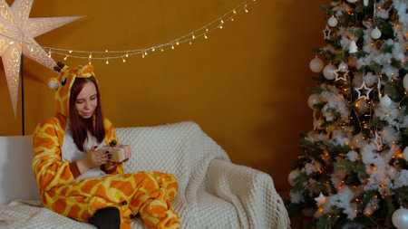 Woman in pajamas with gift box sitting on couch near Christmas tree. Young female considers and shakes christmas present in anticipation. Concept of surprise during holidays.の写真素材