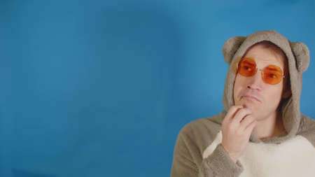 Man in sunglasses and hoodie with ears thoughtfully looks away on blue background with space for text. Pensive male scratches his chin while reflecting about information.の写真素材
