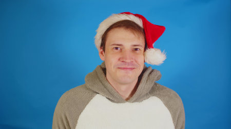 Handsome man in santa hat looking at camera and smiling. Happy male expressing positive emotions on blue background.の写真素材