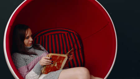 Young woman reads old book, sitting in ball chair. Female resting with shabby book in modern...
