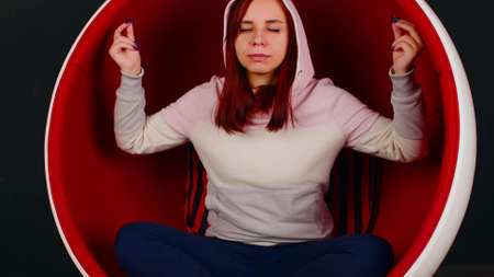 Young woman meditating in ball chair. Young female with closed eyes sitting in butterfly pose with zen gesture while practicing stress relief meditation in ball chair against black backgroundの写真素材