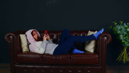 Woman browsing smartphone on couch. Focused female in casual hoodie text messaging on cellphone while resting on comfortable leather sofa in room on black backgroundの写真素材