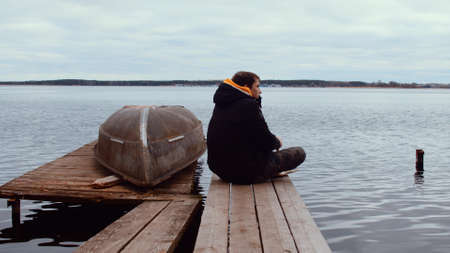 Side view of young man in warm jacket sitting on wooden pier and looking away on background of...