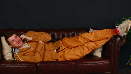 Young man speaks on smartphone, lying on sofa. Handsome male talks on mobile phone, relaxing on...