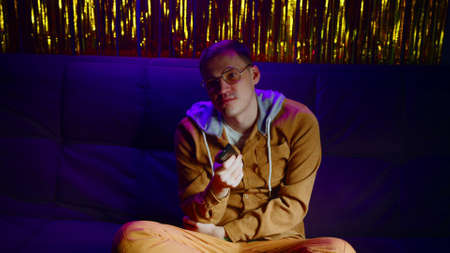 Sad man with remote control sitting on sofa and watching tv in dark room with shimmering light. Dissatisfied male in glasses switches channel watching television without interest in late evening.の写真素材