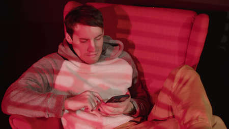 Young man browsing smartphone, sitting in armchair in dark room with falling red light and shadow blinds. Relaxed male using mobile phone.の写真素材