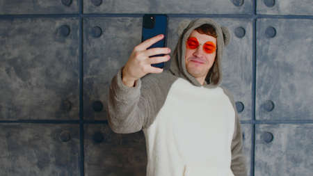 Young man in hood with ears and orange glasses with mobile phone near patterned concrete wall....