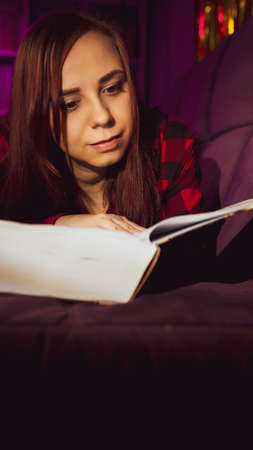 A beautiful student reading a big book. A thoughtful young woman lying on the sofa with a book and reading in a dark room in the eveningの写真素材