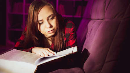 A beautiful student reading a big book. A thoughtful young woman lying on the sofa with a book and reading in a dark room in the eveningの写真素材
