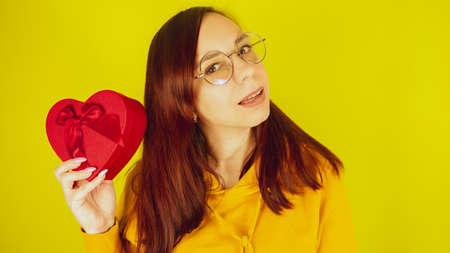Young woman in glasses with red gift box in form of heart on yellow background. Happy female shaking romantic gift and smiling. Concept of present on valentine day and international womens day.の写真素材