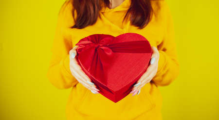 Body part of woman with red gift box in form of heart on yellow background. Unrecognizable person holding romantic gift with ribbon. Concept of present on valentine day and international womens day.の写真素材