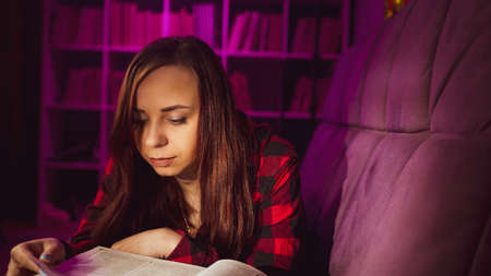 A beautiful student reading a big book. A thoughtful young woman lying on the sofa with a book and...
