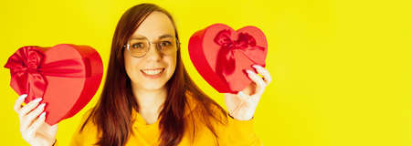 Young woman in glasses with red gift boxes in form of heart on yellow background. Happy female holding romantic gifts, looking at camera and smiling. Concept of presents on international womens day.の写真素材