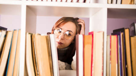 Thoughtful woman in glasses looking at books and scratching head, standing behind bookshelf. Young female reflecting about which book to read. Concept of large selection in library.の写真素材