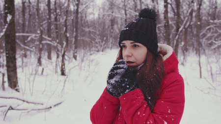 Young frozen woman in in the header and red jacket. Adult female warming hands in gloves with his breath in cold weather.の写真素材