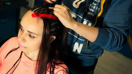 Unrecognizable person makes hairstyle for young woman in salon. Professional hairstylist braids...