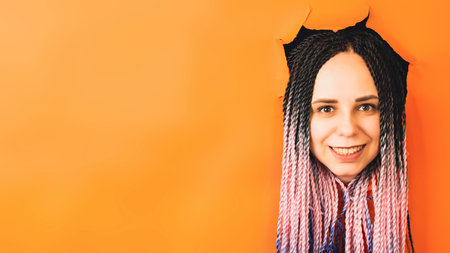 Smiling woman with dreadlocks looking at camera. Cheerful young female with multicolored hair looking at camera in the hole on orange background in studioの写真素材