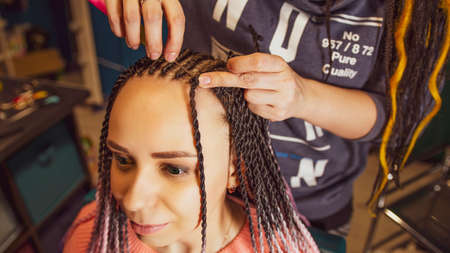 Unrecognizable person makes hairstyle for young woman in salon. Professional hairstylist braids...