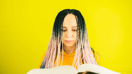 Young woman with senegalese pigtails reading book. Pretty female with multicolored hairstyle...
