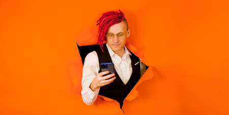 Man with red dreadlocks using smartphone. Businessman with dyed hair looking browsing cellphone whilein the hole in orange background in studioの写真素材