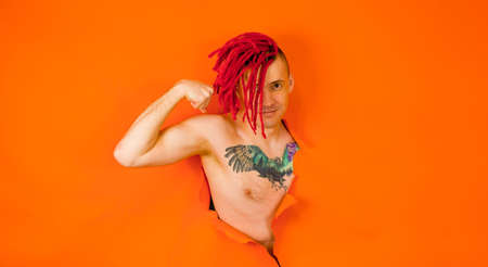 Shirtless man with red dreadlocks and with bird tattoo on chest, showing biceps and looking at...