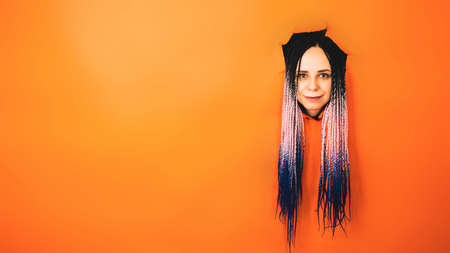 Smiling woman with dreadlocks looking at camera. Cheerful young female with multicolored hair looking at camera in the hole on orange background in studioの写真素材