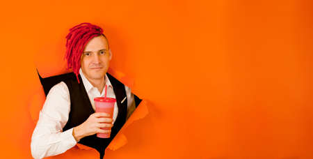 Barman with drink looking through hole in paper background. Male bartender with red dreadlocks and cup of sweet beverage peeking through ripped orange paper background in studio and looking at cameraの写真素材