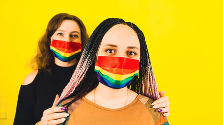 Lesbian couple in rainbow masks on yellow background. Young LGBT women in studio. One female standing in front, other behind and hugs her shoulders.の写真素材