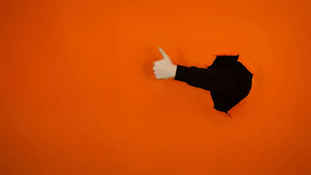 Man's hand showing thumb up and sticking out of hole of orange background. Unrecognizable person...