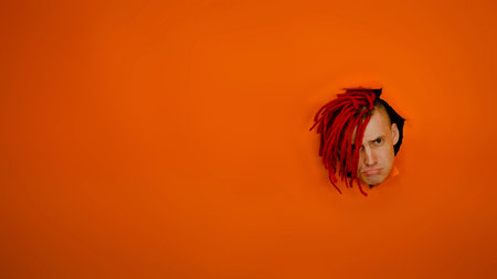 Young man with sad expression on his face sticking out of hole of orange background. Offended male with red dreadlocks purses his lipsの写真素材