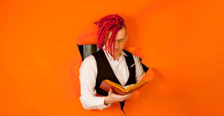 Well dressed man with bright hairstyle reading book and sticking out of hole of orange background....