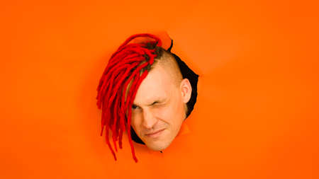 Young man sticking out of hole of orange background. Handsome guy looking at camera and winking