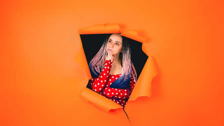 Thoughtful young woman scratching chin and looking out of hole of orange background. Beautiful female looking away and reflecting on lifeの写真素材