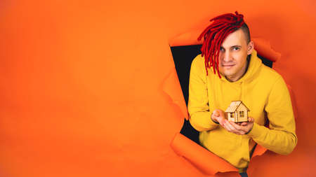 Young handsome man with small wooden house sticking out of hole of orange background. Stylish hipster holding small toy house. Concept of purchasing new apartment and roof overheadの写真素材