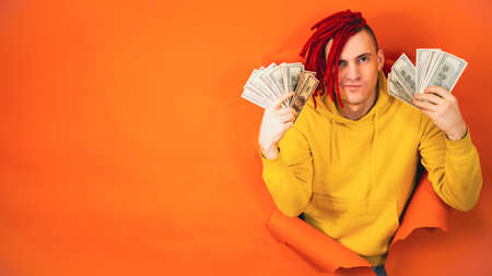 Young handsome man with money sticks out of hole of orange background. Stylish hipster with dollar billsの写真素材