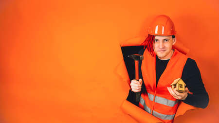 Male construction worker in overalls holds wooden house and sticks out of hole of orange background....