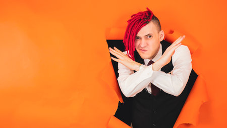 Serious young man with red dreadlocks show stop gesture finish by crossed hands crossing hands, gesturing warning or prohibition, through a hole in the orange wall in the studioの写真素材