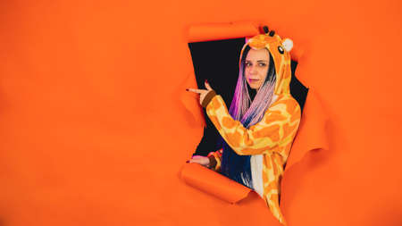 Young woman in pajamas of giraffe sticking out of hole of orange background and pointing index...